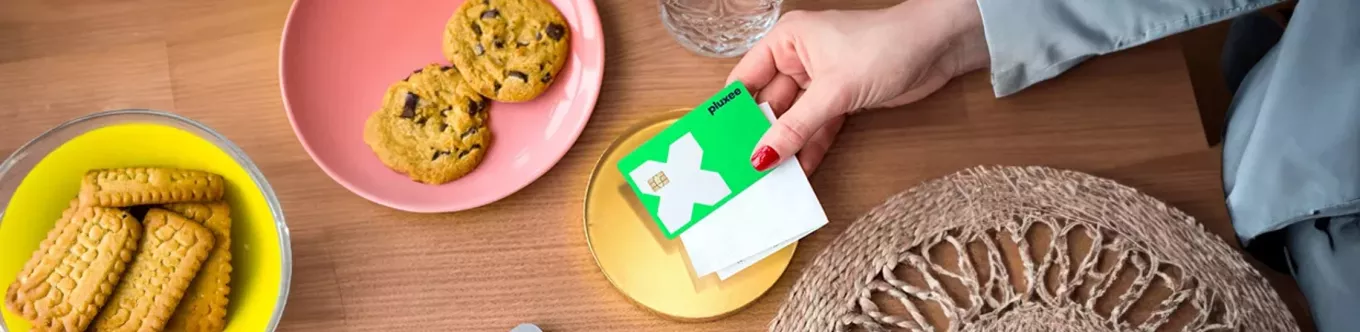 Close-up of a woman's hand placing a green Pluxee card on a receipt on a tray beside cookies at a café, indicating payment. 