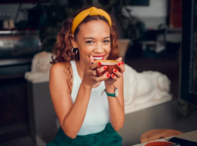 a young woman smiling while holding a cup of coffee up to drink in a coffee shop 