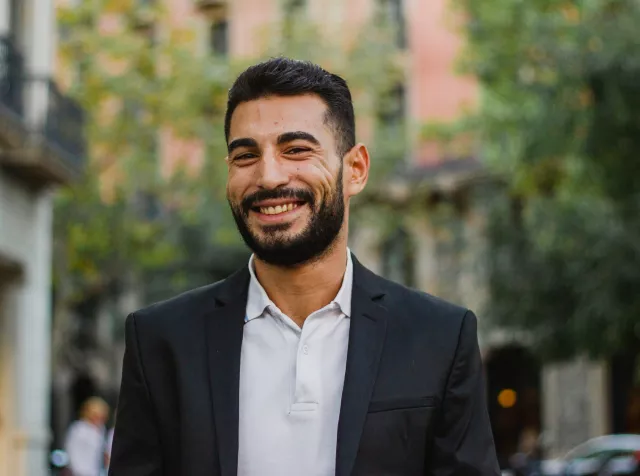 young man in blazer standing on a street with green trees behind him smiling at the camera