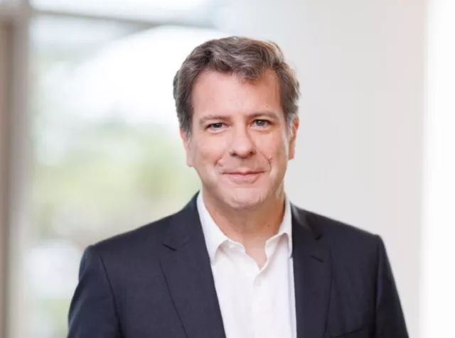 Headshot of Aurelien Sonet​, a white man with grey hair, wearing a light blue tie and white dress shirt, smiling with teeth, and dressed in a dark blue blazer.