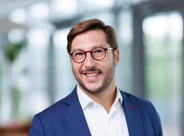 Headshot of Alexandre Cotarmanac’h​, a man with brown hair, wearing red circular glasses, a blue blazer and a white dress shirt smiling with teeth