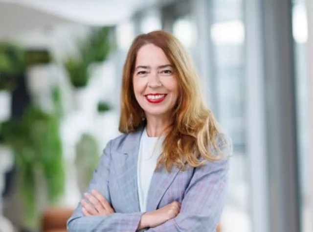 Headshot of Beatrice Bihr, Older woman with long brown hair, wearing a blue and grey checkered blazer and a round blue bead necklace, smiling with teeth and wearing red lipstick.
