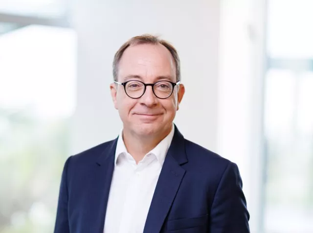 Headshot of Sebastien Godet, an older man with light brown short hair, dressed in a blue blazer and white shirt, and round glasses smiling at the camera