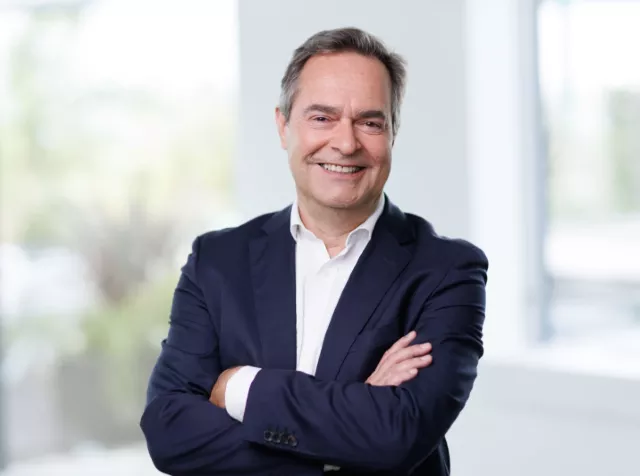 Headshot of Thierry Guihard, an older white man with grey hair wearing a dark blue blazer and white dress shirt, with his arms crossed smiling with teeth