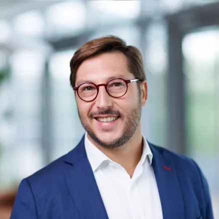 Middle-aged man with brown hair and short stubble, wearing red circular glasses, a blue blazer, and a white dress shirt, smiling at the camera with teeth.