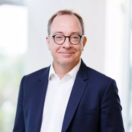 Headshot of Sebastien Godet, an older man with light brown short hair, dressed in a blue blazer and white shirt, and round glasses smiling at the camera