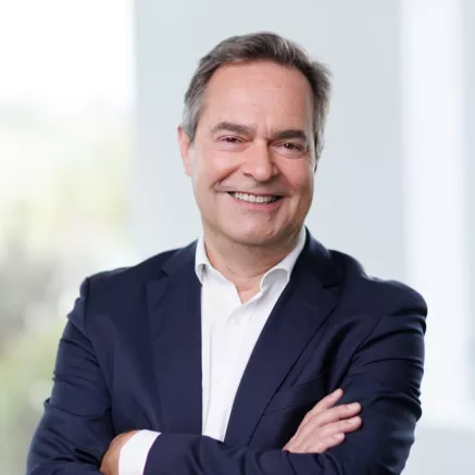 Headshot of Thierry Guihard, an older white man with grey hair wearing a dark blue blazer and white dress shirt, with his arms crossed smiling with teeth